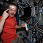 Hadfield is wearing an orange polo shirt and khaki pants and holding a silver canister with a white label and blue tip in his left hand. With his right hand, he is pointing to a round yellow sensor taped to his forehead. There is a laptop over his left shoulder and multiple cords, wires, and switches on the wall in front of him.